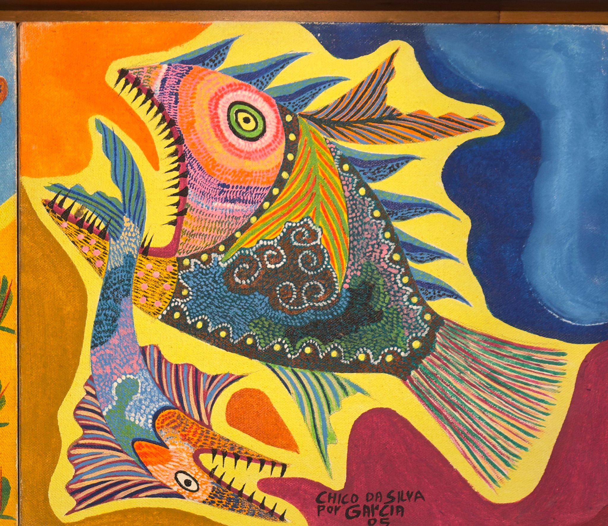 Peixe grande de cabeça rosa e olho verde, boca aberta de dentes pretos, espinhos azuis e cauda listrada, na tela de cima, à direita. Detalhe de Animais Fantásticos, Chico da Silva por Garcia, 2005–2006.