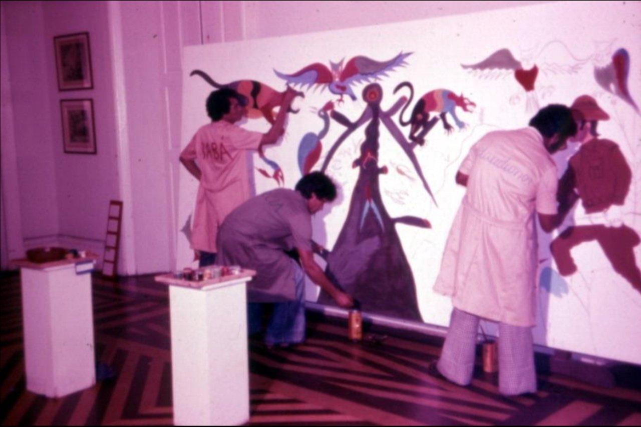 Três homens de jaleco pintam um painel grande de bichos coloridos sobre fundo branco: à esquerda, um de pé, de costas, com BABA escrito nas costas; no meio, outro agachado junto à base; à direita, um terceiro de pé. No chão de tacos, pedestais brancos com latas de tinta. Foto em cores desbotadas.
