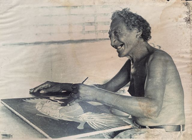Homem idoso e grisalho, sem camisa, ri de perfil enquanto se curva sobre uma prancha escura posta na mesa, a mão esquerda segurando o papel e a direita conduzindo um instrumento fino sobre uma ave de asas abertas, desenhada em branco sobre o fundo escuro. Reprodução em preto e branco frio.