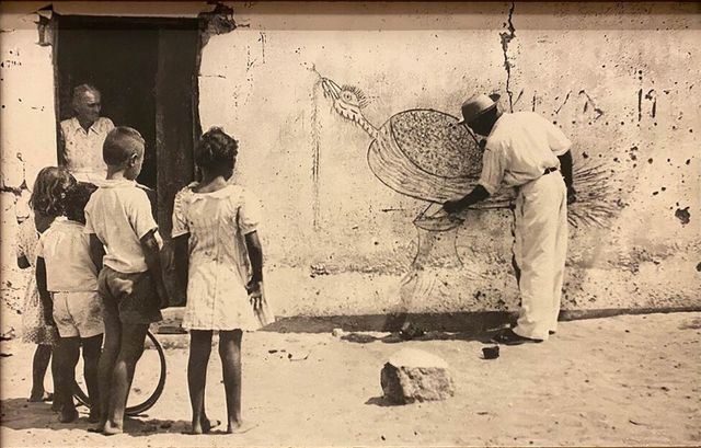On a sandy street, four barefoot children stand with their backs to the camera, watching a man in a hat and light clothes who bends over a large long-tailed bird drawn on the mud wall of a house. A man leans in the dark doorway; a bicycle rim and a white stone lie on the ground. Black and white photograph.