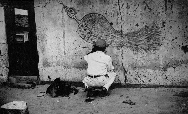 A man in a hat, crouching with his back to the camera, draws a long-tailed bird with a large eye and fanned plumage on the mud wall of a house. Black and white photograph.