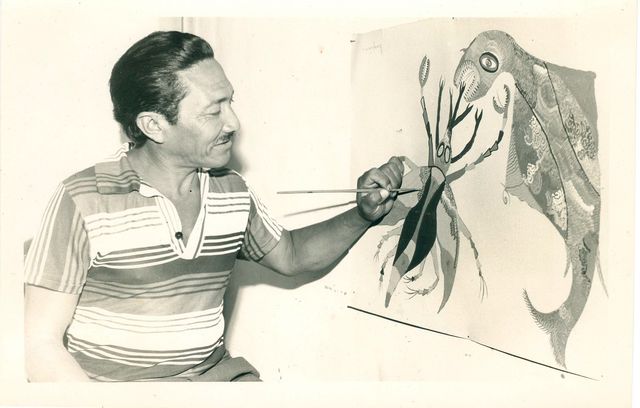 A man in a striped polo shirt, seen in profile, holds a fine brush to the leg of a large creature painted in gouache: a curved beak, a round eye, a segmented crab-like body and a wing with a fan-shaped, patterned tail. The sheet rests against a plain wall. Black and white photograph.