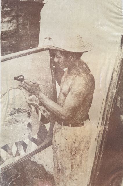 A bare-chested man in a straw hat paints with a fine brush on a canvas propped on a rough wooden easel, in a narrow gap between the canvas and a plank wall. On the canvas, a fish with triangular teeth already fills the lower half. Sepia-toned reproduction with visible halftone screen.