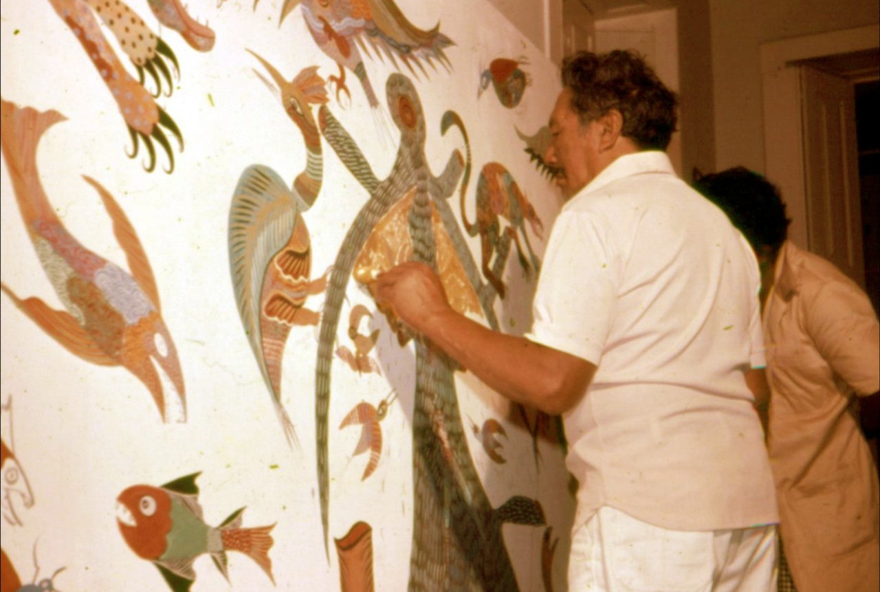 Homem de camisa e calça brancas, visto de costas, encosta a mão numa área dourada de um painel grande: um lagarto comprido e listrado no eixo, uma ave azul de asas em leque, um peixe vermelho e verde embaixo, tudo sobre fundo branco. Atrás dele há outra pessoa; à direita, o batente de uma porta. Fotografia em cores.