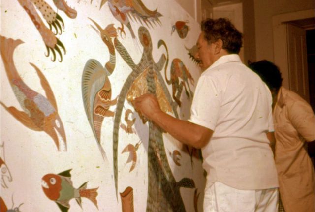 Homem de camisa e calça brancas, visto de costas, encosta a mão numa área dourada de um painel grande: um lagarto comprido e listrado no eixo, uma ave azul de asas em leque, um peixe vermelho e verde embaixo, tudo sobre fundo branco. Atrás dele há outra pessoa; à direita, o batente de uma porta. Fotografia em cores.