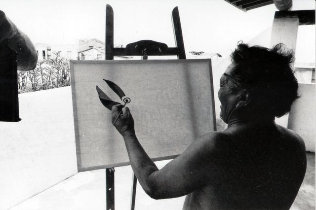 A shirtless man with his hair tied back, seen from behind, paints a canvas on a wooden easel outdoors; only two dark leaves and a round eye exist on it so far. Black and white photograph, low houses in the background.