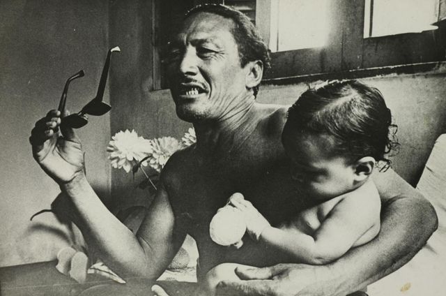 A shirtless man laughing, one arm raised holding a pair of sunglasses; against his chest a small child sleeps. Black and white photograph, indoors, with a window and a vase of flowers.