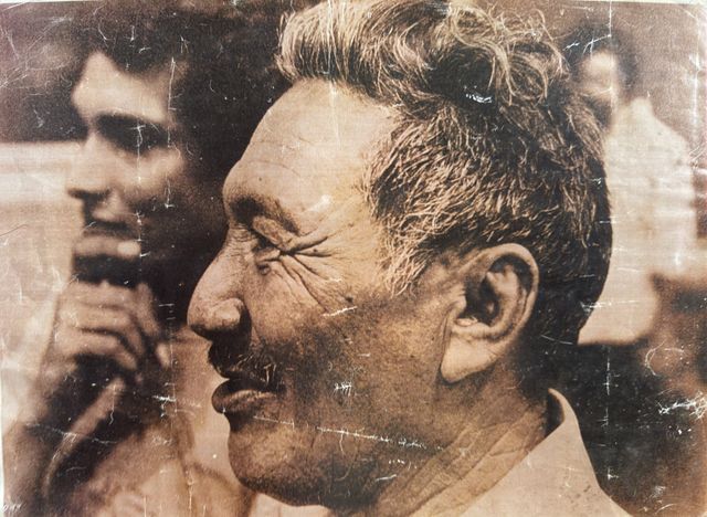 Close profile of an older man with white hair, mouth slightly open as if speaking, deep creases at the corner of the eye. Behind him, out of focus, a young man with his hand near his face and two other figures. Sepia-toned reproduction with visible halftone screen.