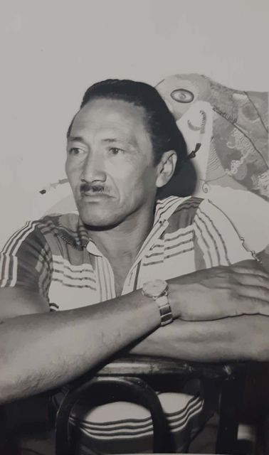 A man in a striped polo shirt sits astride a wooden chair, both arms folded over the backrest, a metal-strapped watch on his wrist. On the wall behind him hangs a painting of a round-eyed fantastic creature. Black and white photograph.
