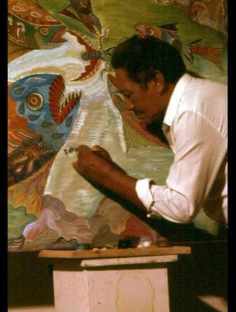 Homem de camisa branca, de perfil, escreve com um pincel fino sobre a faixa clara de um painel pintado com peixes, aves e folhagens em verde, azul e vermelho.