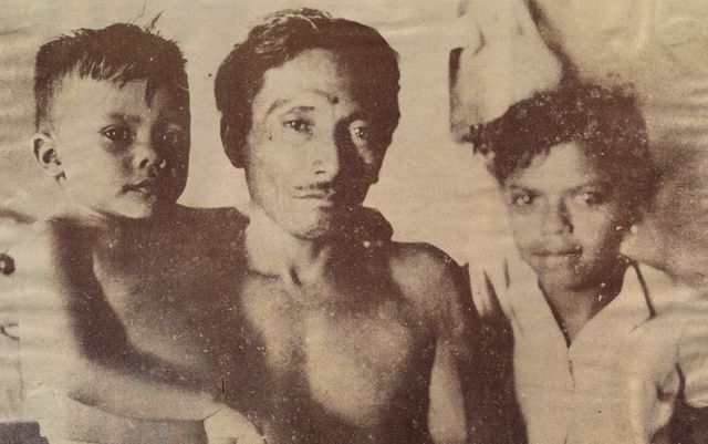 A young man with a thin moustache, bare-chested, looks at the camera between two children: on the left a small boy with spiky hair, on the right a girl in a light blouse with her hand on his shoulder. Sepia-toned reproduction with visible halftone screen.