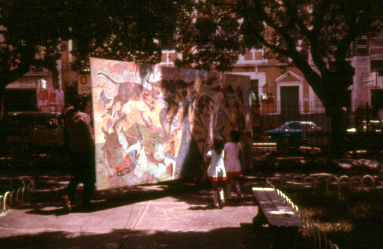 Sob a sombra de árvores grandes, um painel pintado, erguido de pé, é levado por uma praça; um homem o segura pela esquerda, e duas meninas de vestido claro olham de perto. Ao fundo, um carro azul e sobrados.