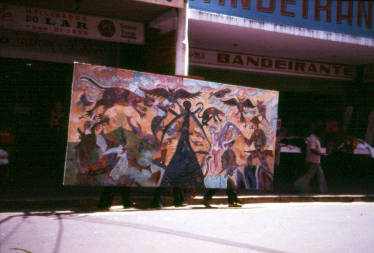 Um painel largo, tomado de bichos em vermelho, azul, ocre e preto, atravessa a calçada de pé, carregado por pessoas escondidas atrás dele; só as pernas aparecem por baixo. Ao fundo, fachadas de lojas, uma com o letreiro BANDEIRANTE.