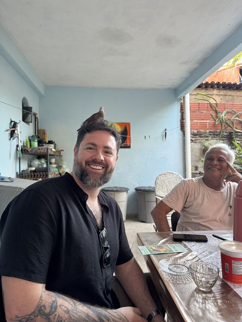 Um homem de barba grisalha sorri para a câmera com um pequeno pássaro cinza equilibrado no alto da cabeça; ao fundo, um homem mais velho ri sentado a uma mesa com xícaras de café.