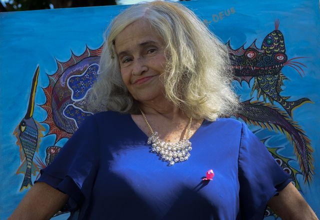 An older woman with loose white hair and a dark blue blouse stands smiling in the sun before a large blue canvas of fantastic fish, one of them dark with red eye-like discs.